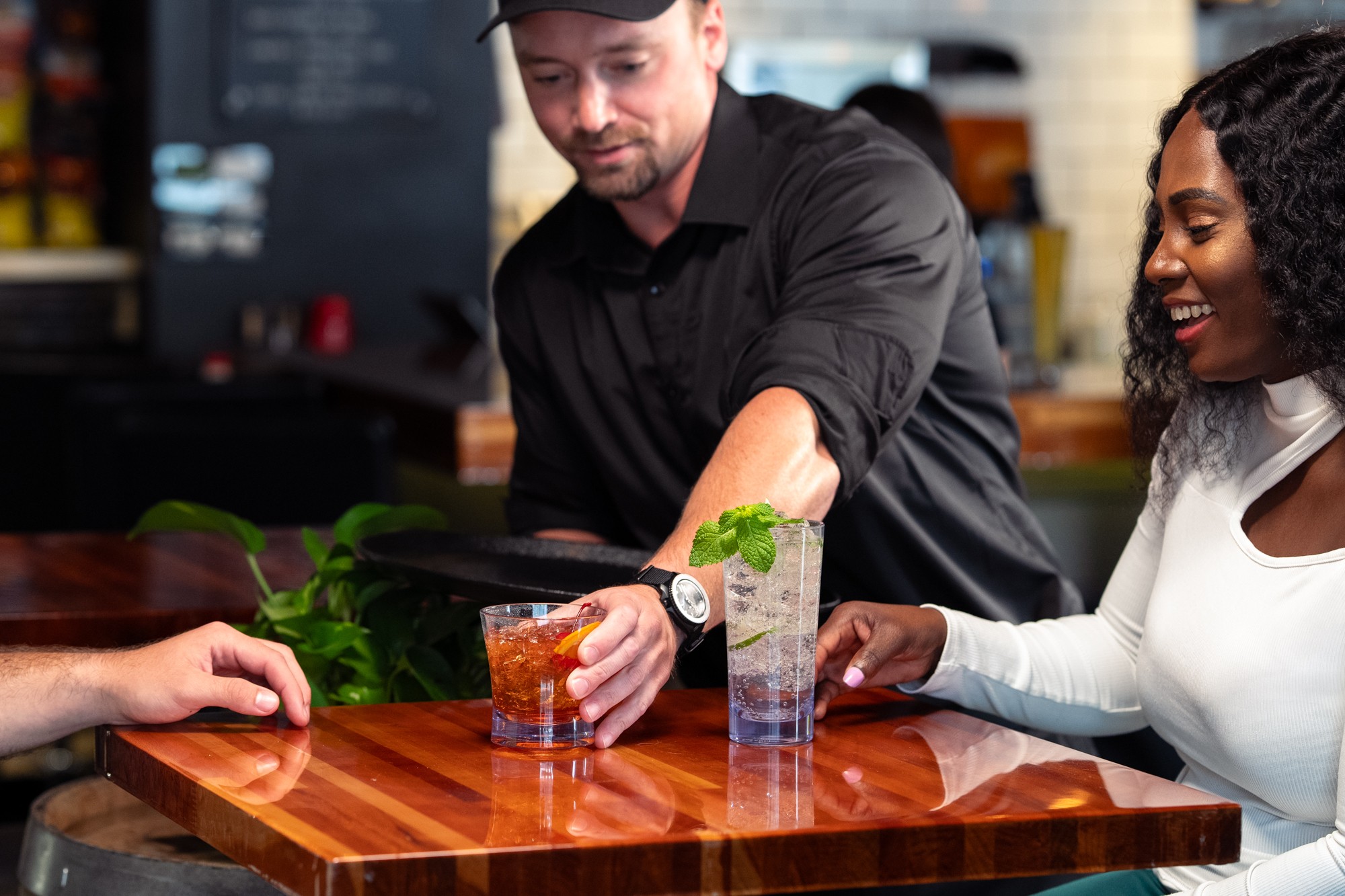 Patron enjoying a cocktail at a restaurant as a server delivers another cocktail to the table.
